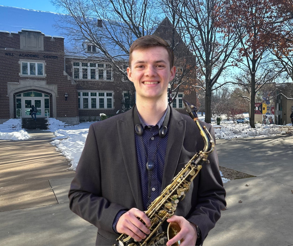 Eli Regier in a brown suit jacket and dark blue shirt, holding his saxophone, standing on the Bethel campus with Will Academic Center in the background