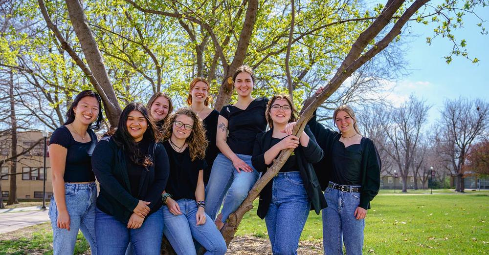 Photo of Woven, eight women in blue jeans and black shirts standing under a tree