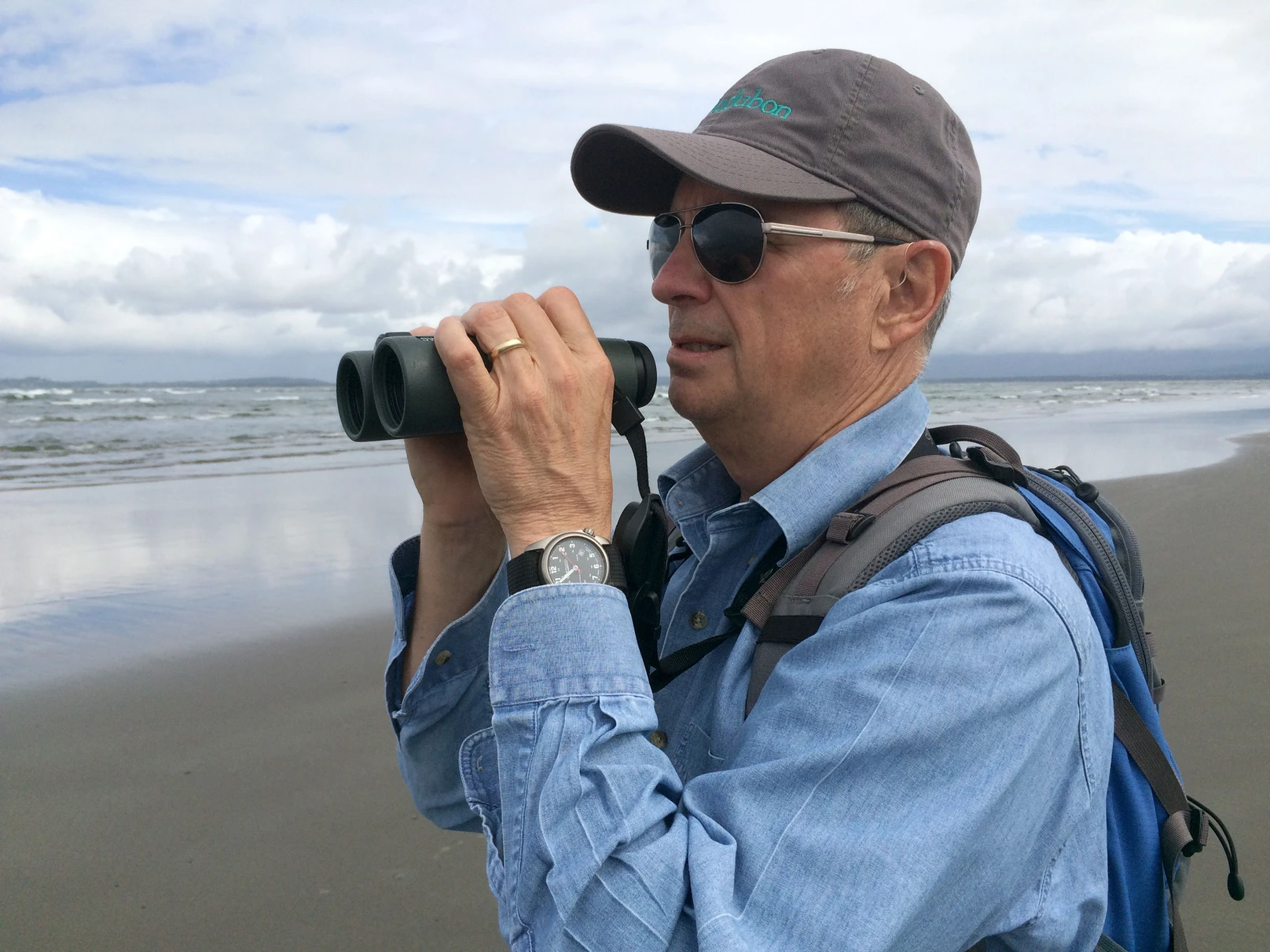 Stan Senner, wearing a ball cap, sunglasses and denim shirt, holding binoculars and standing by the ocean