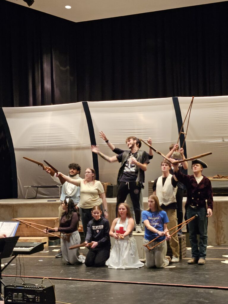 Rehearsal photo showing a group of students on stage, singing, some of them waving rifles (props)