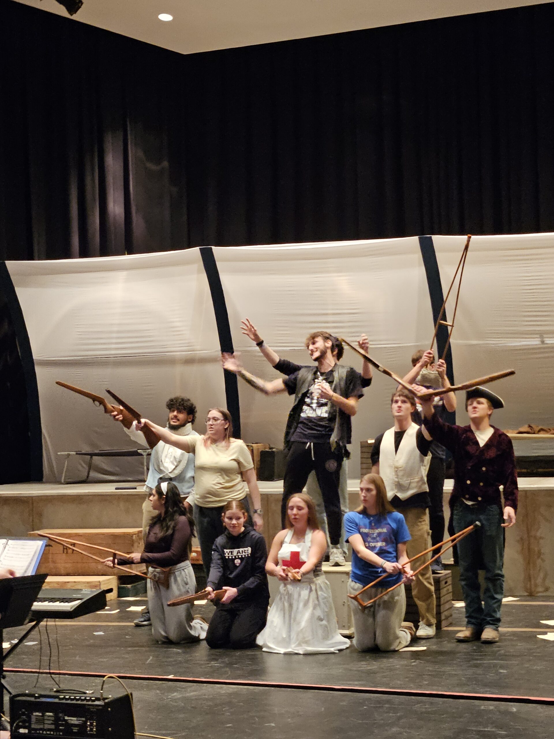 Rehearsal photo showing a group of students on stage, singing, some of them waving rifles (props)
