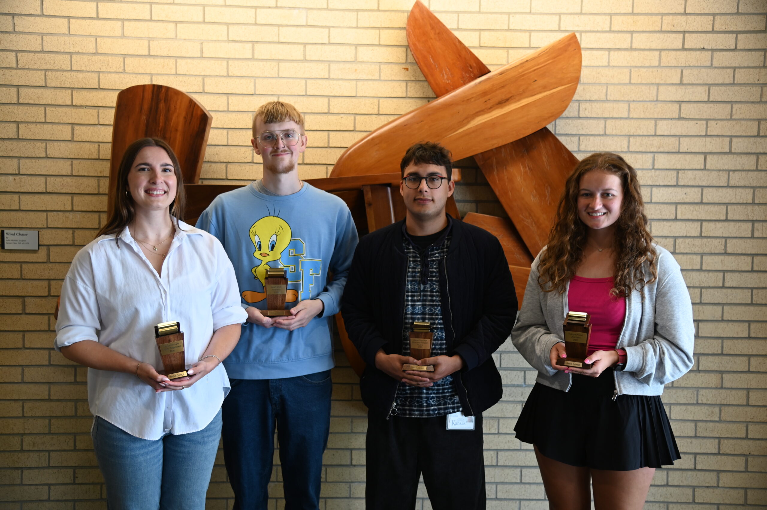 The four Thresher Award winners from 2026 standing in a row in front of a wooden sculpture hanging on a blond brick wall