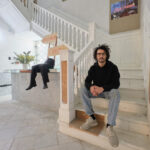 Rodrigo Cacho, a young Latinx man with dark curly hair wearing a black shirt, gray pants and glasses, sitting at the bottom of a long white staircase.