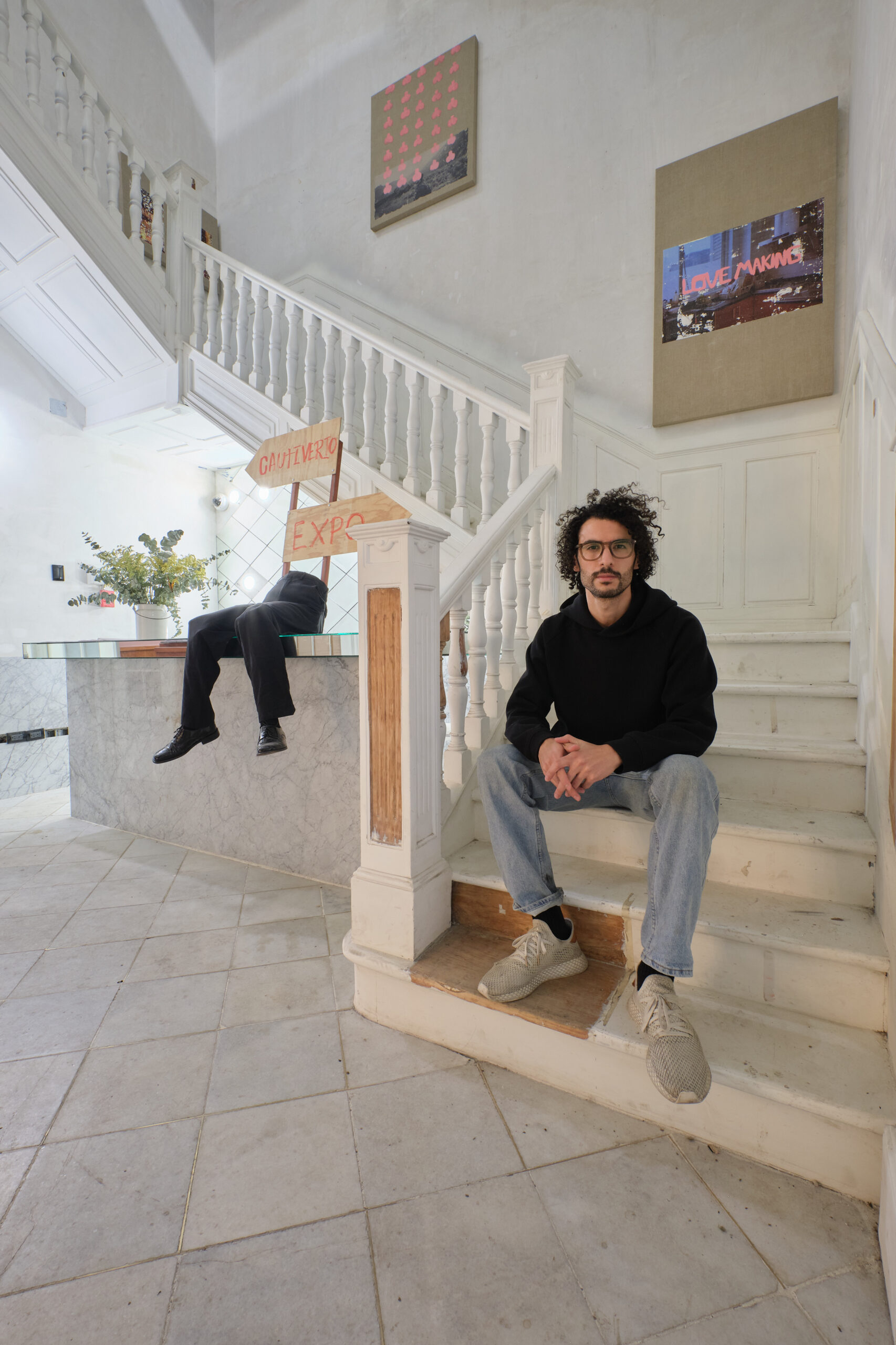Rodrigo Cacho, a young Latinx man with dark curly hair wearing a black shirt, gray pants and glasses, sitting at the bottom of a long white staircase.