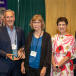 Five people in a line at an awards ceremony; second from left is holding the award.
